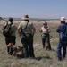 A group of approximately six people, including what appear to be National Park Service rangers in uniform, are gathered on an elevated grassy overlook in a badlands landscape. A female ranger appears to be briefing or leading a discussion with the group, which includes a dog on a leash. The terrain in the background features characteristic eroded buttes and rolling grasslands consistent with the Great Plains badlands region.