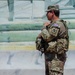A U.S. Army soldier in MultiCam (OCP) camouflage uniform stands at attention or on guard duty near what appears to be a large marble or stone structure. The soldier is wearing a tactical vest with pouches, carrying a radio, and has a name tape partially visible reading 'ROPE...' on their patrol cap. A U.S. flag patch and a unit patch featuring an 'X' design are visible on the left sleeve.