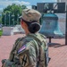 A female U.S. Army soldier in Operational Camouflage Pattern (OCP) uniform stands facing a large bronze bell replica, likely a Liberty Bell replica, at an outdoor memorial or public space. The soldier wears a name tape reading 'VALLEJOS' and displays a unit patch on her left sleeve along with a U.S. flag patch. The setting appears to be Washington, D.C., based on the architecture, brick plaza, and surrounding monuments visible in the background.