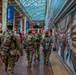 Three National Guard soldiers in camouflage uniforms are patrolling the interior of what appears to be Washington Union Station, identifiable by Amtrak signage and a Hudson store advertisement. The soldiers are walking away from the camera through a busy public transit concourse with a glass-and-steel arched ceiling. The scene suggests a security presence deployment at a major transportation hub.
