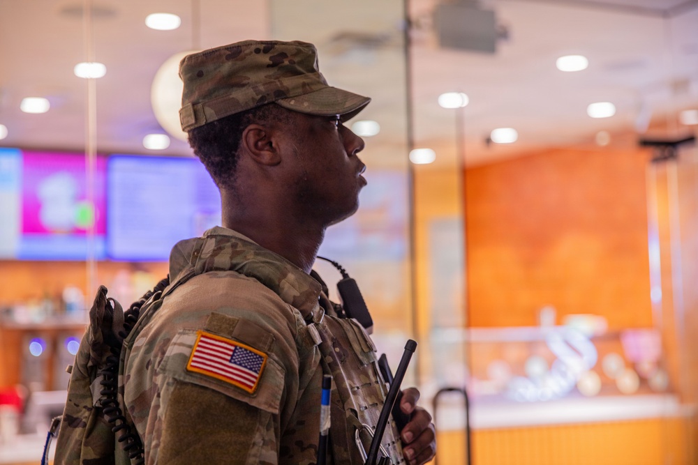 A U.S. Army soldier in Operational Camouflage Pattern (OCP) uniform stands in profile view inside what appears to be a commercial or retail establishment. The soldier is equipped with tactical gear, a radio/communication device, and bears a U.S. flag patch on the left shoulder. The background shows display screens and a glass-enclosed area consistent with a mall, food court, or similar civilian venue.