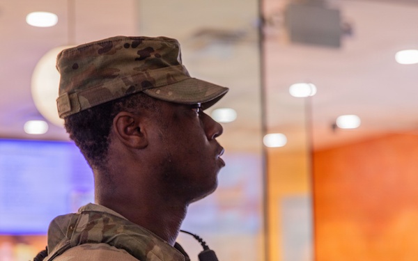 A U.S. Army soldier in Operational Camouflage Pattern (OCP) uniform stands in profile view inside what appears to be a commercial or retail establishment. The soldier is equipped with tactical gear, a radio/communication device, and bears a U.S. flag patch on the left shoulder. The background shows display screens and a glass-enclosed area consistent with a mall, food court, or similar civilian venue.