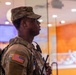 A U.S. Army soldier in Operational Camouflage Pattern (OCP) uniform stands in profile view inside what appears to be a commercial or retail establishment. The soldier is equipped with tactical gear, a radio/communication device, and bears a U.S. flag patch on the left shoulder. The background shows display screens and a glass-enclosed area consistent with a mall, food court, or similar civilian venue.