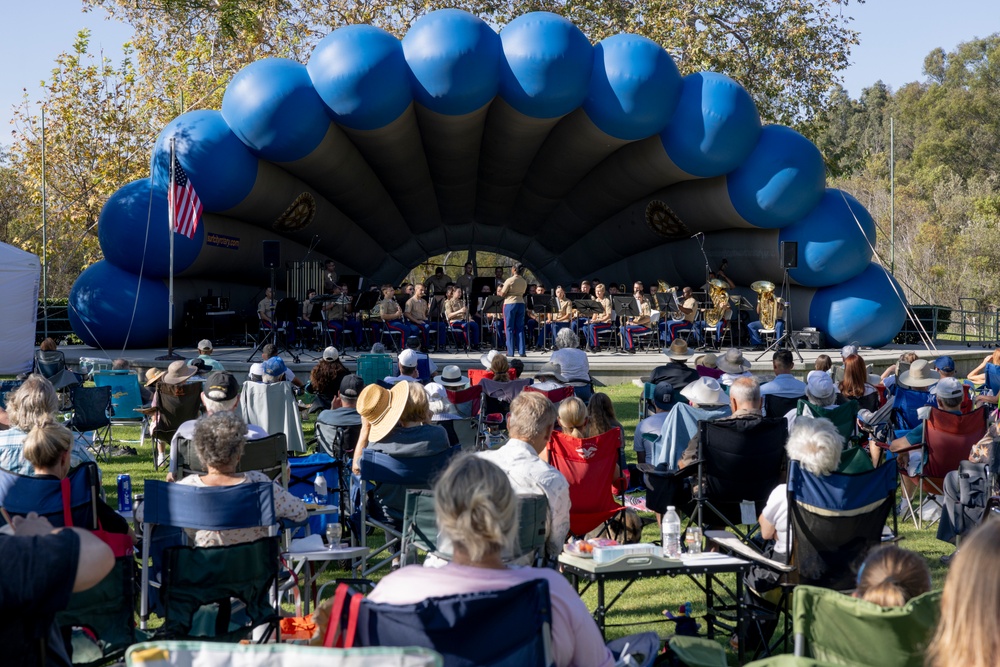 An outdoor military band concert is taking place in a park setting, with a large ensemble performing on a covered stage framed by a large blue inflatable arch. A conductor stands at the front of the uniformed musicians, while a sizeable audience watches from personal lawn chairs and folding seats on the grass. An American flag is visible on the stage, suggesting a patriotic or community event.