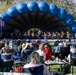 An outdoor military band concert is taking place in a park setting, with a large ensemble performing on a covered stage framed by a large blue inflatable arch. A conductor stands at the front of the uniformed musicians, while a sizeable audience watches from personal lawn chairs and folding seats on the grass. An American flag is visible on the stage, suggesting a patriotic or community event.