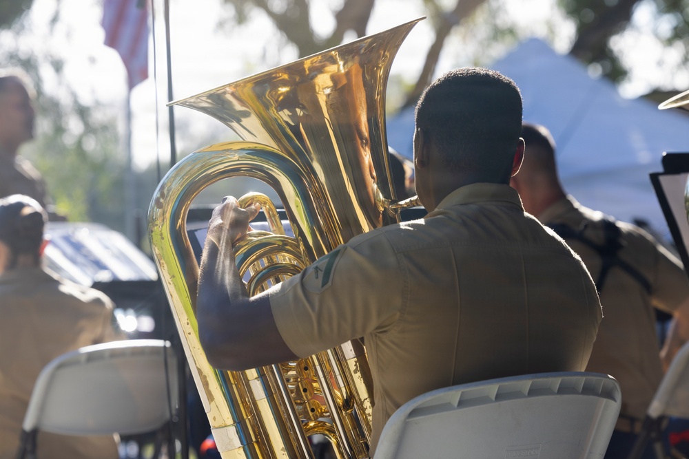 A musician in a tan/khaki uniform is seated and playing a large brass tuba at what appears to be an outdoor event or ceremony. The image is taken from behind the player, showing the large bell of the instrument prominently. Other musicians and attendees are visible in the blurred background.