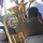 3rd MAW Band Performs at Huntington Beach A musician in a tan/khaki uniform is seated and playing a large brass tuba at what appears to be an outdoor event or ceremony. The image is taken from behind the player, showing the large bell of the instrument prominently. Other musicians and attendees are visible in the blurred background.