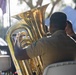 A musician in a tan/khaki uniform is seated and playing a large brass tuba at what appears to be an outdoor event or ceremony. The image is taken from behind the player, showing the large bell of the instrument prominently. Other musicians and attendees are visible in the blurred background.