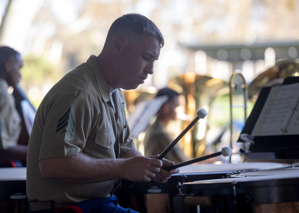 A U.S. Marine Corps sergeant is playing percussion instruments, likely timpani or concert drums, during an outdoor military band performance. He is wearing the standard tan/khaki service uniform with sergeant chevrons visible on his left sleeve. Other band members and music stands with sheet music are visible in the blurred background.