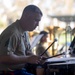 A U.S. Marine Corps sergeant is playing percussion instruments, likely timpani or concert drums, during an outdoor military band performance. He is wearing the standard tan/khaki service uniform with sergeant chevrons visible on his left sleeve. Other band members and music stands with sheet music are visible in the blurred background.