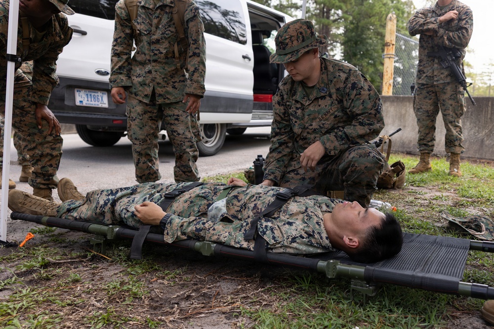 Military personnel in MARPAT camouflage uniforms are conducting a casualty care or medical training exercise. One service member is strapped to a litter/stretcher on the ground while another kneels beside them providing simulated medical attention. Additional personnel observe the exercise near a white Ford Transit van in an outdoor setting.