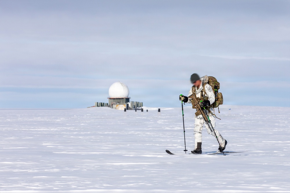 A military or special operations personnel in white winter camouflage is cross-country skiing across a vast snow-covered flat terrain while carrying a tactical backpack and what appears to be a weapon. In the background, a radar dome (radome) installation is visible, suggesting a remote Arctic or Antarctic military/research facility. The subject's face appears to have been deliberately blurred, likely for operational security reasons.