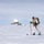 Green Berets, Danish SOF Ski Across Greenland Ice Cap During Operation Apex Indigo A military or special operations personnel in white winter camouflage is cross-country skiing across a vast snow-covered flat terrain while carrying a tactical backpack and what appears to be a weapon. In the background, a radar dome (radome) installation is visible, suggesting a remote Arctic or Antarctic military/research facility. The subject's face appears to have been deliberately blurred, likely for operational security reasons.
