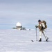 A military or special operations personnel in white winter camouflage is cross-country skiing across a vast snow-covered flat terrain while carrying a tactical backpack and what appears to be a weapon. In the background, a radar dome (radome) installation is visible, suggesting a remote Arctic or Antarctic military/research facility. The subject's face appears to have been deliberately blurred, likely for operational security reasons.