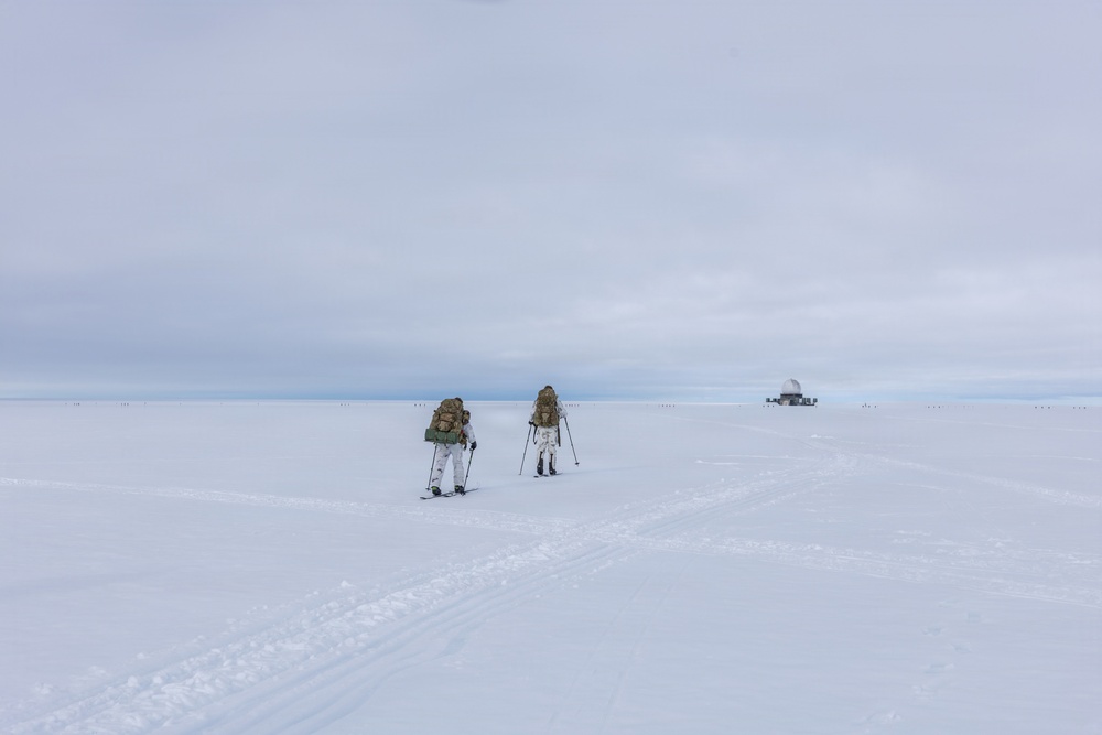 Two individuals in white winter camouflage gear are cross-country skiing across a vast, flat snow-covered landscape while carrying large military-style backpacks and using ski poles. In the far background to the right, a white dome-shaped structure — likely a radar or communications installation — is visible on the horizon. The scene suggests a military arctic or polar training exercise.