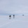 Green Berets, Danish SOF Ski Across Greenland Ice Cap During Operation Apex Indigo Two individuals in white winter camouflage gear are cross-country skiing across a vast, flat snow-covered landscape while carrying large military-style backpacks and using ski poles. In the far background to the right, a white dome-shaped structure — likely a radar or communications installation — is visible on the horizon. The scene suggests a military arctic or polar training exercise.
