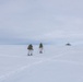 Two individuals in white winter camouflage gear are cross-country skiing across a vast, flat snow-covered landscape while carrying large military-style backpacks and using ski poles. In the far background to the right, a white dome-shaped structure — likely a radar or communications installation — is visible on the horizon. The scene suggests a military arctic or polar training exercise.