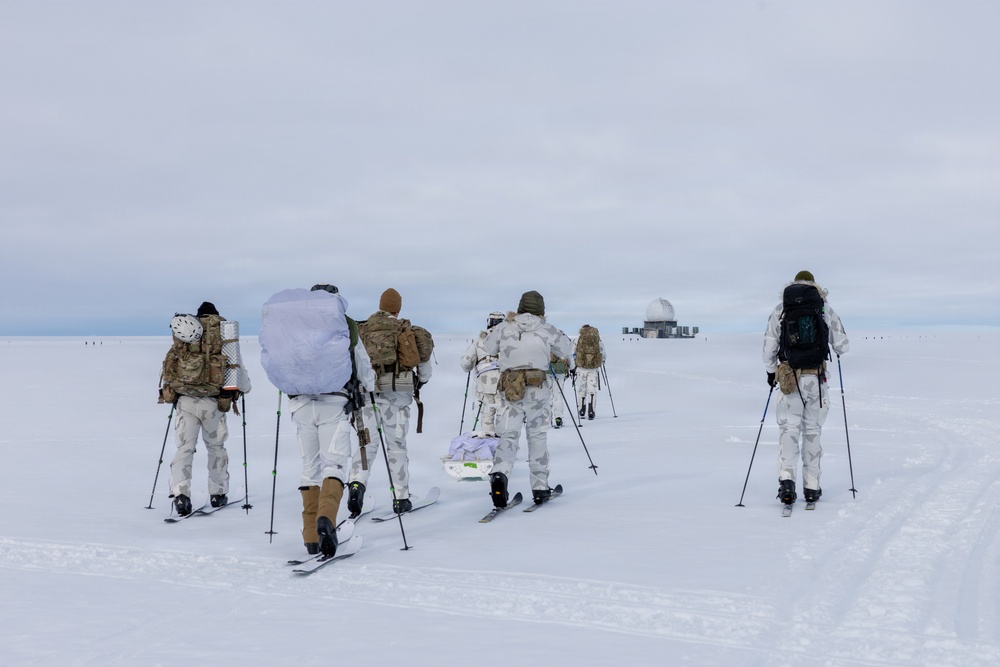 A group of military personnel in winter camouflage uniforms are cross-country skiing across a snow-covered arctic or polar landscape. They are carrying heavy tactical backpacks and equipment, moving toward what appears to be a remote installation or outpost visible in the background. The scene suggests a military arctic warfare training exercise or operational patrol.
