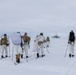 A group of military personnel in winter camouflage uniforms are cross-country skiing across a snow-covered arctic or polar landscape. They are carrying heavy tactical backpacks and equipment, moving toward what appears to be a remote installation or outpost visible in the background. The scene suggests a military arctic warfare training exercise or operational patrol.