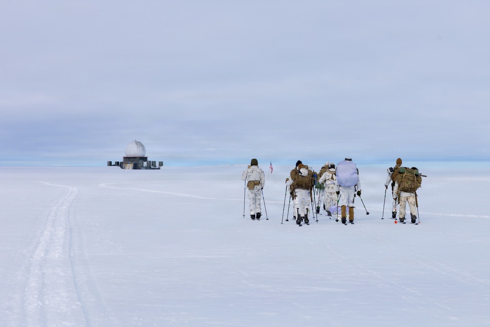 A group of approximately six individuals in winter camouflage gear and military-style equipment are gathered on a vast, flat snow-covered landscape. They are carrying ski poles, large backpacks, and at least one American flag is visible among the group. In the background to the left, a distant structure with a dome — likely a radar or communications installation — is visible on the horizon.