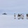 Green Berets, Danish SOF Ski Across Greenland Ice Cap During Operation Apex Indigo A group of approximately six individuals in winter camouflage gear and military-style equipment are gathered on a vast, flat snow-covered landscape. They are carrying ski poles, large backpacks, and at least one American flag is visible among the group. In the background to the left, a distant structure with a dome — likely a radar or communications installation — is visible on the horizon.