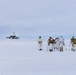 A group of approximately six individuals in winter camouflage gear and military-style equipment are gathered on a vast, flat snow-covered landscape. They are carrying ski poles, large backpacks, and at least one American flag is visible among the group. In the background to the left, a distant structure with a dome — likely a radar or communications installation — is visible on the horizon.
