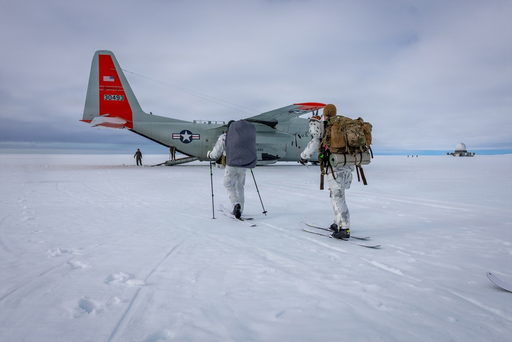 Two personnel in white winter camouflage gear and skis approach a U.S. military LC-130 Hercules aircraft (tail number 30493) equipped with ski-wheel landing gear on a snow/ice surface. The aircraft bears U.S. Air National Guard markings with distinctive red-orange wing and tail accents. A dome-shaped structure is visible in the far background, suggesting a polar research or military installation.