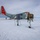 Green Berets, Danish SOF Ski Across Greenland Ice Cap During Operation Apex Indigo Two personnel in white winter camouflage gear and skis approach a U.S. military LC-130 Hercules aircraft (tail number 30493) equipped with ski-wheel landing gear on a snow/ice surface. The aircraft bears U.S. Air National Guard markings with distinctive red-orange wing and tail accents. A dome-shaped structure is visible in the far background, suggesting a polar research or military installation.