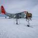Two personnel in white winter camouflage gear and skis approach a U.S. military LC-130 Hercules aircraft (tail number 30493) equipped with ski-wheel landing gear on a snow/ice surface. The aircraft bears U.S. Air National Guard markings with distinctive red-orange wing and tail accents. A dome-shaped structure is visible in the far background, suggesting a polar research or military installation.