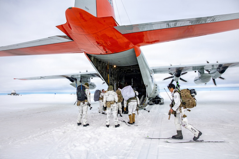 A group of military personnel in white winter camouflage uniforms are boarding or loading equipment onto a large cargo aircraft with a distinctive orange and white paint scheme, consistent with polar/Antarctic operations support aircraft. The scene takes place on a snow and ice surface, suggesting an Arctic or Antarctic environment. Personnel are carrying heavy backpacks and at least one individual is equipped with skis.