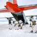 A group of military personnel in white winter camouflage uniforms are boarding or loading equipment onto a large cargo aircraft with a distinctive orange and white paint scheme, consistent with polar/Antarctic operations support aircraft. The scene takes place on a snow and ice surface, suggesting an Arctic or Antarctic environment. Personnel are carrying heavy backpacks and at least one individual is equipped with skis.