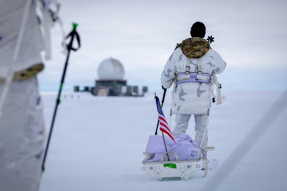 A person in white winter camouflage military gear and a multicam jacket is pulling a sled across a snow-covered landscape. An American flag is planted on the sled, which carries bundled supplies. In the background, a radar dome and military installation structures are visible on the horizon.