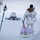 Green Berets, Danish SOF Ski Across Greenland Ice Cap During Operation Apex Indigo A person in white winter camouflage military gear and a multicam jacket is pulling a sled across a snow-covered landscape. An American flag is planted on the sled, which carries bundled supplies. In the background, a radar dome and military installation structures are visible on the horizon.