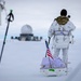A person in white winter camouflage military gear and a multicam jacket is pulling a sled across a snow-covered landscape. An American flag is planted on the sled, which carries bundled supplies. In the background, a radar dome and military installation structures are visible on the horizon.