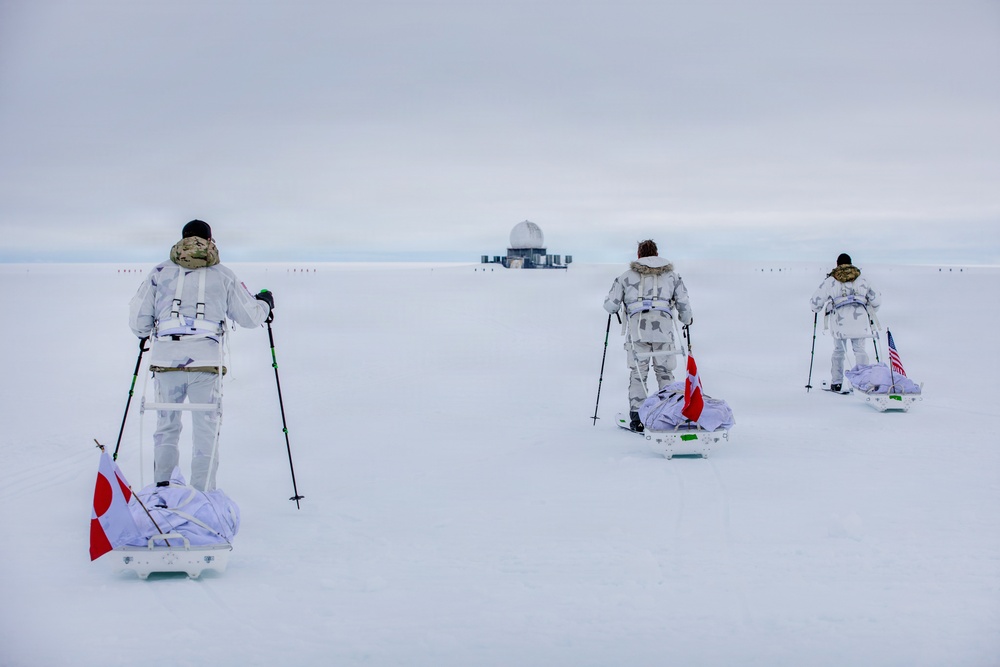 Three individuals in white winter camouflage gear are skiing across a snow-covered arctic or polar landscape, each pulling a sled (pulk) behind them. They are approaching a distant facility or installation visible on the horizon. Each person's sled displays a different national flag: Greenland, Canada, and the United States.