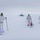 Green Berets, Danish SOF Ski Across Greenland Ice Cap During Operation Apex Indigo Three individuals in white winter camouflage gear are skiing across a snow-covered arctic or polar landscape, each pulling a sled (pulk) behind them. They are approaching a distant facility or installation visible on the horizon. Each person's sled displays a different national flag: Greenland, Canada, and the United States.