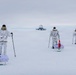 Three individuals in white winter camouflage gear are skiing across a snow-covered arctic or polar landscape, each pulling a sled (pulk) behind them. They are approaching a distant facility or installation visible on the horizon. Each person's sled displays a different national flag: Greenland, Canada, and the United States.