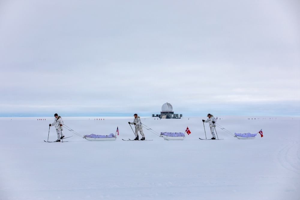 Three individuals in white winter camouflage gear are skiing across a vast snow-covered landscape while pulling pulk sleds. Each sled displays a national flag, including a visible American flag and Danish flag. In the background, a domed structure consistent with a polar research station or radar installation is visible on the horizon.