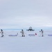 Three individuals in white winter camouflage gear are skiing across a vast snow-covered landscape while pulling pulk sleds. Each sled displays a national flag, including a visible American flag and Danish flag. In the background, a domed structure consistent with a polar research station or radar installation is visible on the horizon.