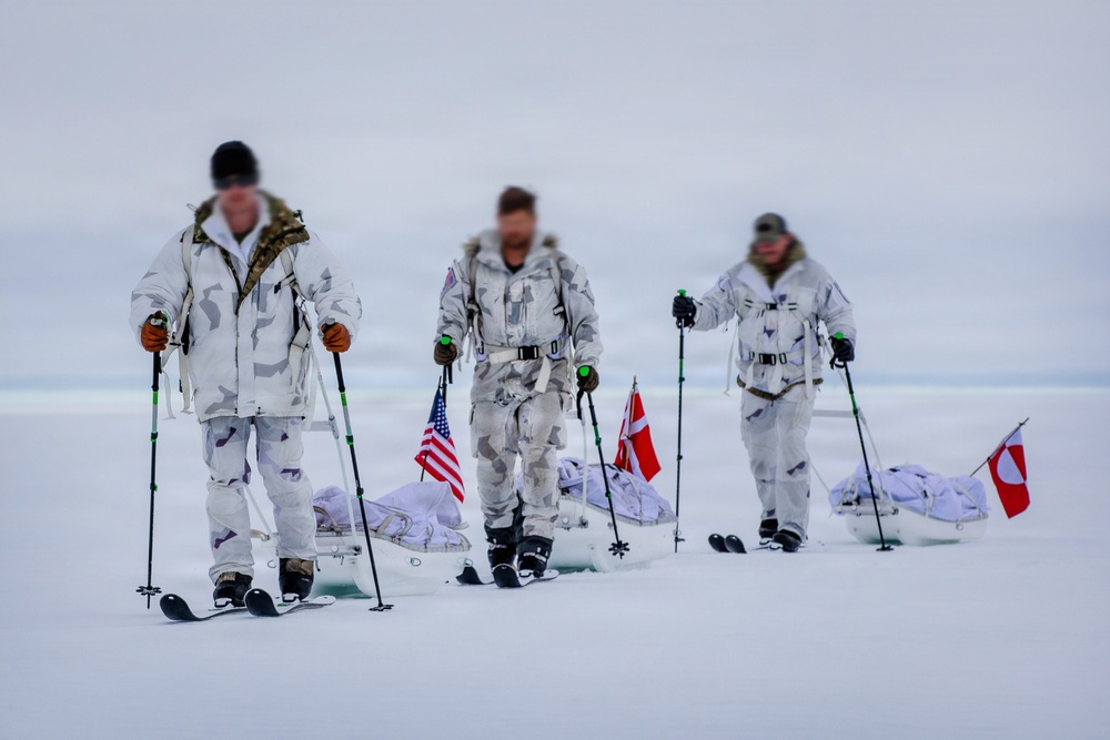 Three individuals in white winter camouflage uniforms are skiing across a snow-covered arctic or polar landscape while pulling sleds (pulkas). They are carrying ski poles and display three flags: an American flag, a Danish flag, and what appears to be the flag of Greenland. The scene suggests a joint military or expedition operation in an extreme cold-weather environment.