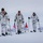 Green Berets, Danish SOF Ski Across Greenland Ice Cap During Operation Apex Indigo Three individuals in white winter camouflage uniforms are skiing across a snow-covered arctic or polar landscape while pulling sleds (pulkas). They are carrying ski poles and display three flags: an American flag, a Danish flag, and what appears to be the flag of Greenland. The scene suggests a joint military or expedition operation in an extreme cold-weather environment.