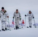 Three individuals in white winter camouflage uniforms are skiing across a snow-covered arctic or polar landscape while pulling sleds (pulkas). They are carrying ski poles and display three flags: an American flag, a Danish flag, and what appears to be the flag of Greenland. The scene suggests a joint military or expedition operation in an extreme cold-weather environment.
