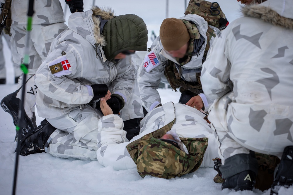 Military personnel in white winter camouflage uniforms are conducting a combat casualty care or medical training exercise in a snowy environment. At least one person is lying on the ground while two or more individuals attend to them, suggesting a simulated trauma or first aid scenario. Patches visible on the uniforms indicate the presence of both Danish and American military personnel.