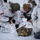 Green Berets, Danish SOF Ski Across Greenland Ice Cap During Operation Apex Indigo Military personnel in white winter camouflage uniforms are conducting a combat casualty care or medical training exercise in a snowy environment. At least one person is lying on the ground while two or more individuals attend to them, suggesting a simulated trauma or first aid scenario. Patches visible on the uniforms indicate the presence of both Danish and American military personnel.