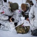 Military personnel in white winter camouflage uniforms are conducting a combat casualty care or medical training exercise in a snowy environment. At least one person is lying on the ground while two or more individuals attend to them, suggesting a simulated trauma or first aid scenario. Patches visible on the uniforms indicate the presence of both Danish and American military personnel.