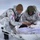 Green Berets, Danish SOF Ski Across Greenland Ice Cap During Operation Apex Indigo Two military personnel in white arctic camouflage uniforms are attending to a casualty lying on a white rescue sled/litter in a snow-covered environment. One individual wears a US flag patch and a red cross medical patch, while the other wears a Danish flag patch, indicating a joint multinational military medical exercise or operation. They appear to be performing field medical treatment in extreme cold-weather conditions.