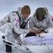 Two military personnel in white arctic camouflage uniforms are attending to a casualty lying on a white rescue sled/litter in a snow-covered environment. One individual wears a US flag patch and a red cross medical patch, while the other wears a Danish flag patch, indicating a joint multinational military medical exercise or operation. They appear to be performing field medical treatment in extreme cold-weather conditions.