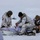 Green Berets, Danish SOF Ski Across Greenland Ice Cap During Operation Apex Indigo Three military personnel in white arctic camouflage uniforms are conducting what appears to be a field medical exercise or casualty treatment on a snow-covered terrain. Two identifiable national flags are visible — Danish (red and white cross) and American (stars and stripes) — suggesting a joint multinational Arctic operation. A person is lying on a white sled/pulk while the others attend to them with a visible medical kit.