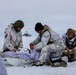 Three military personnel in white arctic camouflage uniforms are conducting what appears to be a field medical exercise or casualty treatment on a snow-covered terrain. Two identifiable national flags are visible — Danish (red and white cross) and American (stars and stripes) — suggesting a joint multinational Arctic operation. A person is lying on a white sled/pulk while the others attend to them with a visible medical kit.