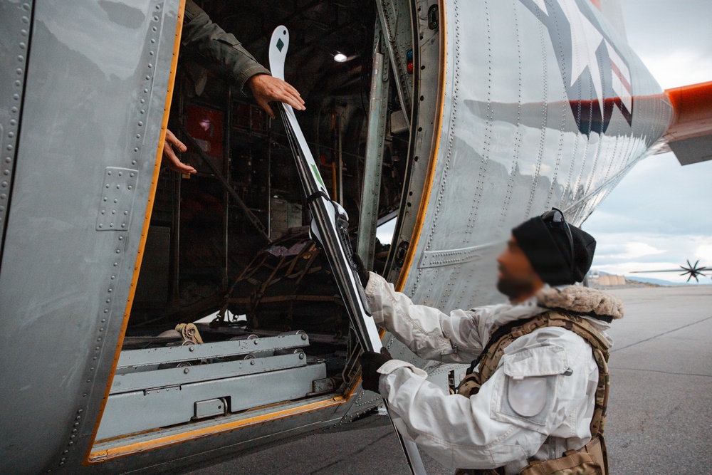 A person in a white winter suit and tactical vest is loading or unloading ski equipment into/from a military-style helicopter. The individual's face has been deliberately blurred, suggesting operational security or privacy concerns. The scene appears to take place on a tarmac or airfield in an overcast environment.